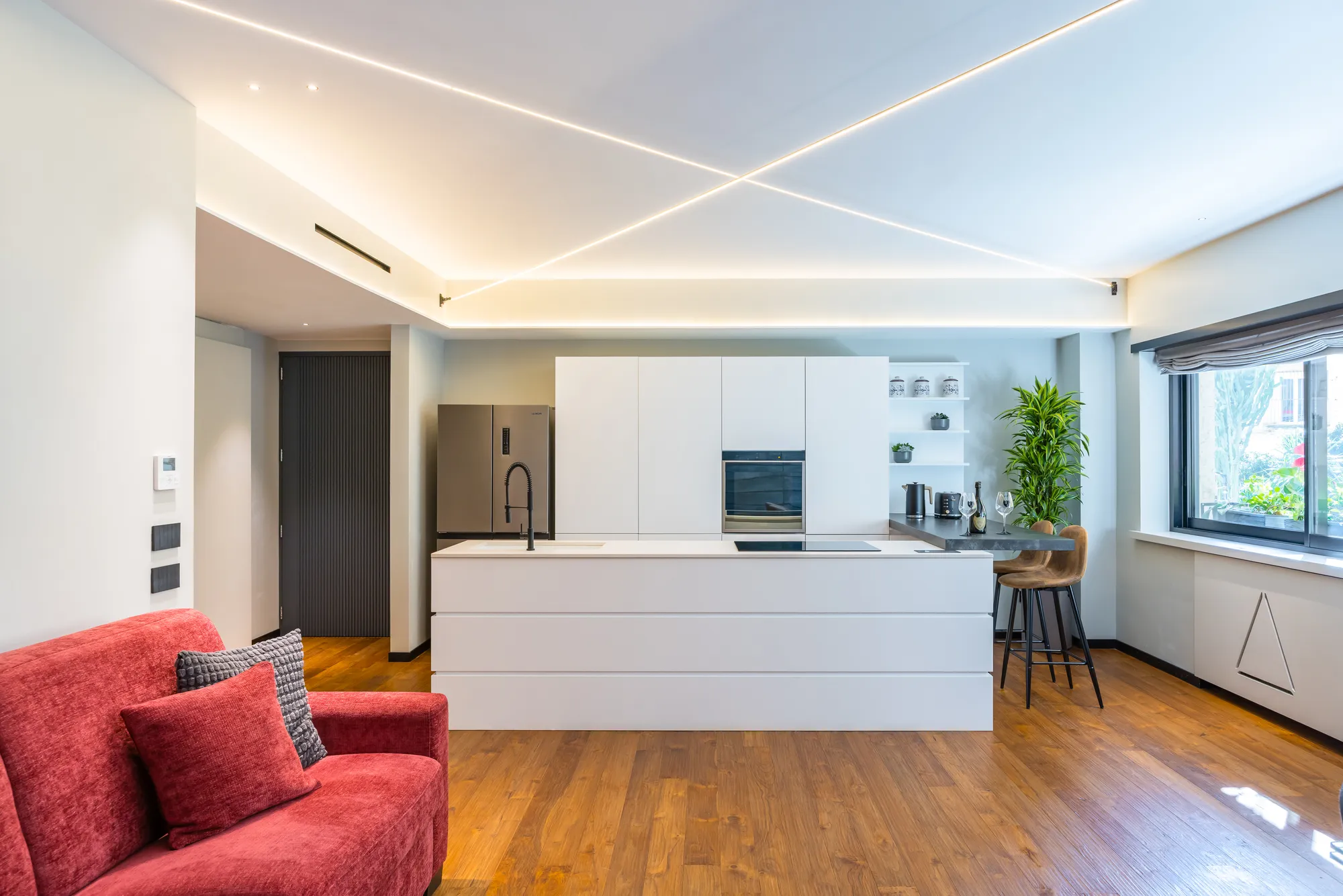 Salone di Santopaolo 1, cucina a isola bianca, divano rosso e sgabelli sotto le linee di luce a soffitto